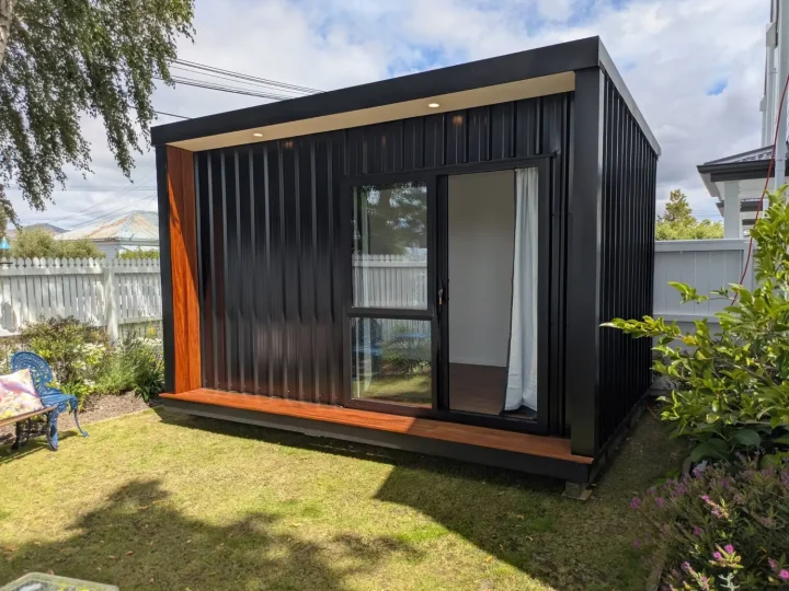 Black cabin with a kwila deck and reveal on a garden lawn beside a white picket fence