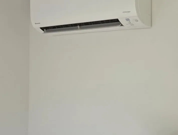A heat pump mounted high on the wall of the treatment-room cabin, with a smoke alarm on the ceiling