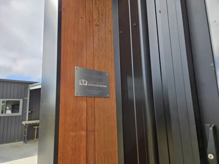Kwila board carrying the Cube Cabins maker's plaque beside black steel cladding
