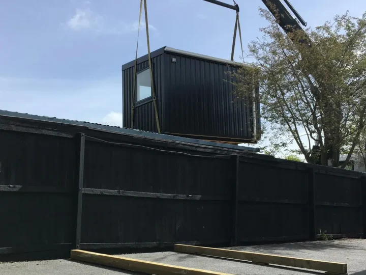 A black cabin lifted over a tall fence by crane