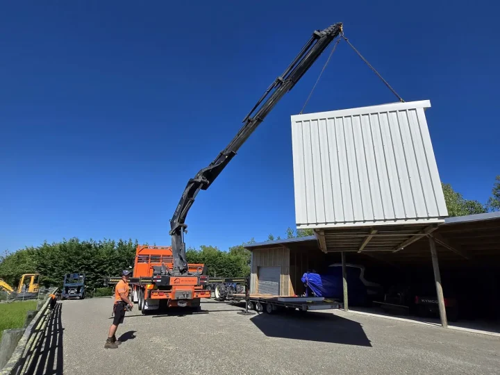 A white cabin lifted off a truck by crane beside a farm shed